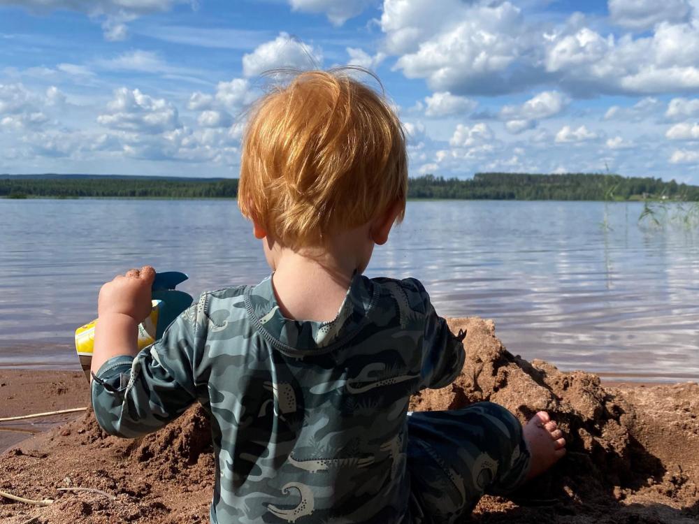 En liten pojke som sitter ner i sanden och har sjön framför sig. 
