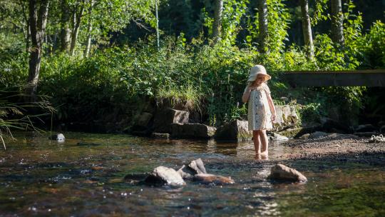 En liten flicka i gul klänning står barfota vid kanten till en å.